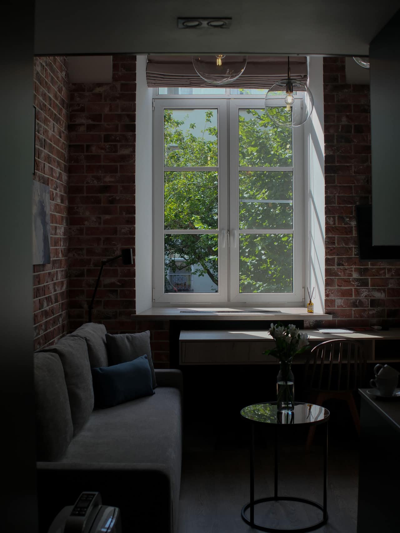 Salón en penumbra: una ventana grande que da a un árbol, pared de ladrillo visto y una lámpara colgando del techo.